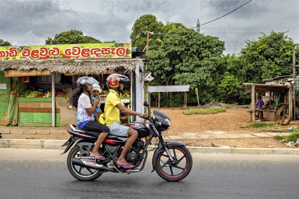Two people on a motor scooter ride down a street, past a small shop and tropical plants, a family on a moped in the streets of Colombo Sri Lanka