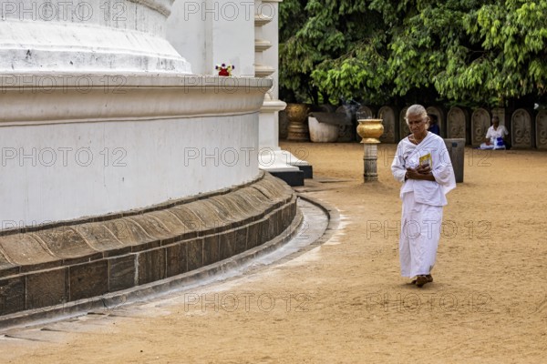 A man in meditative posture walks around a large white temple with a book, People praying at Kelaniya Temple in Colombo Sri Lanka