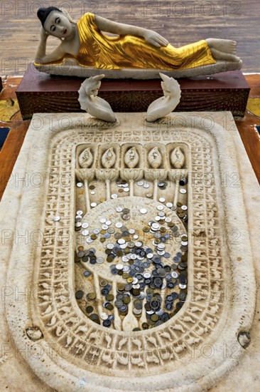 A reclining Buddha statue with gold robes above a carved stone surface filled with coins, symbolizing offerings in a spiritual setting, the Seema Malaka temple with the Buddha statues in Colombo Sri Lanka