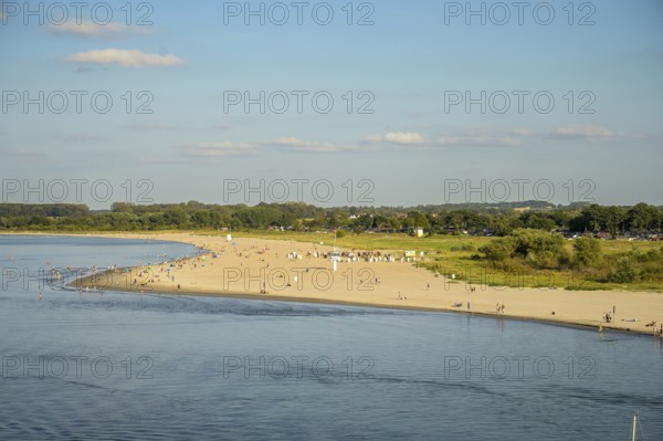 A sunny beach on Priwall near Rosenhagen with people on the shore, surrounded by green countryside and calm sea taken by the Scandinavian ferry entering Travemünde harbour, Lübeck-Travemünde, Schleswig-Holstein, Germany