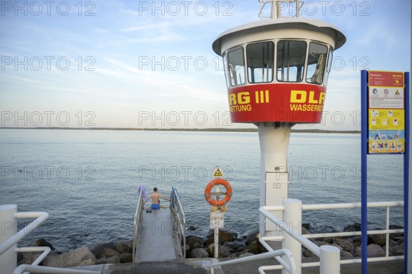 A modern DLRG watchtower looks out at the vast water while a bather sits on the jetty, Lübeck-Travemünde, Schleswig-Holstein, Germany