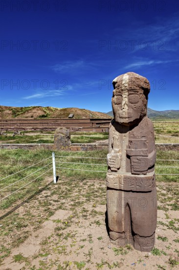 Free-standing stone statue in a grass-covered ruined landscape under a blue sky, The archaeological site with the ruins of Tiwanaku in Bolivia