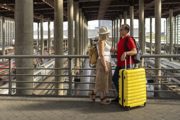 Middle aged couple enjoying a travel moment at a busy train station while waiting for their train. They stand together with luggage and look happy and relaxed. Image represents tourism, modern lifestyle, mobility and travel experiences