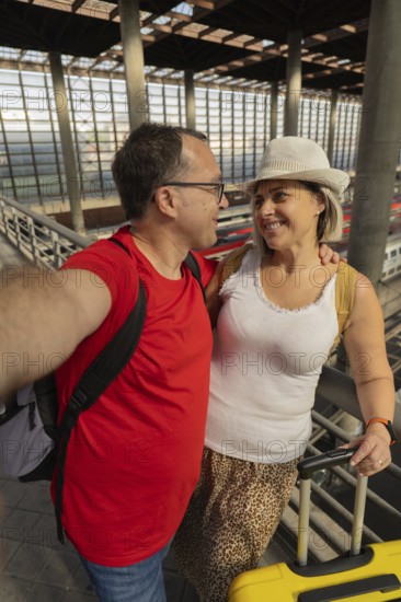 Smiling adult travel couple taking a selfie while looking at each other inside a modern train station with luggage. Romantic travel moment, connection, tourism and lifestyle concept