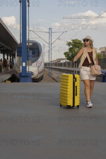 Middle aged woman pulling a bright yellow suitcase on a modern urban train platform, enjoying a calm travel moment while waiting for public transportation