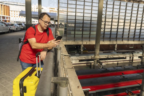 Solo male traveler waiting at a modern railway station, holding a yellow suitcase and checking his phone while looking over the train platforms below