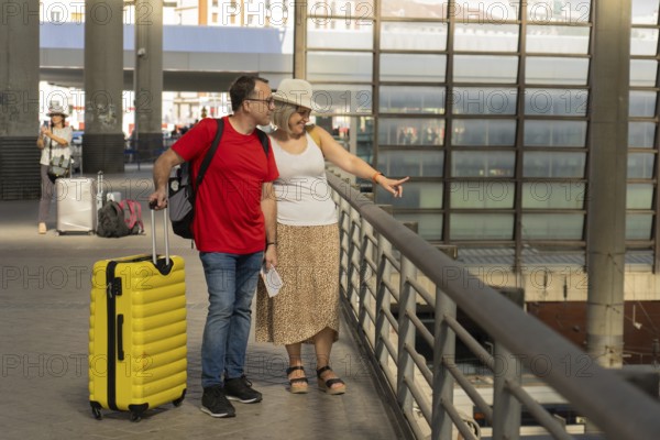 Adult travel couple walking together inside a modern transportation terminal above train tracks, pulling luggage while talking and pointing. Multimodal transport, travel connection, tourism and urban mobility concept