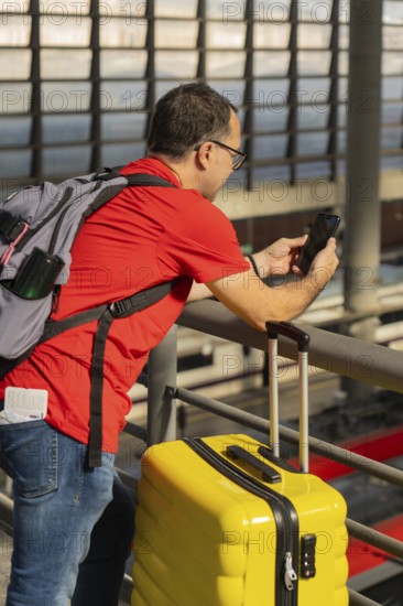 Mature male traveler using a smartphone at a train station while standing next to a yellow suitcase. Concept of travel, technology, urban mobility and modern tourism