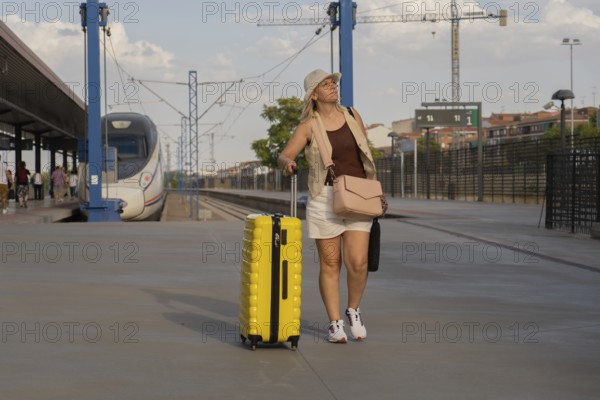 Adult woman walking along a modern train station platform, pulling a yellow suitcase while waiting for her train during a relaxed urban travel moment