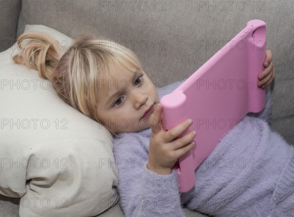 Portrait of a blonde girl, 4 years old, lying down reading a tablet in Ystad, Skåne County, Sweden, Scandinavia