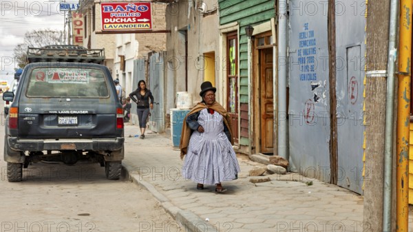 Woman wearing traditional clothes wearing melon walks on a sidewalk with colorful houses and murals, people in an altiplano near Uyuni in Bolivia