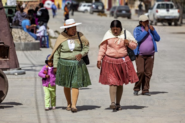 Two woman and a little girl walk down a busy street in traditional clothes, people in the altiplano near Uyuni in Bolivia