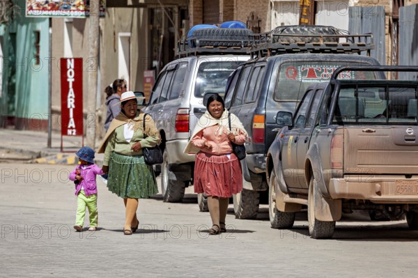 Two woman and a child in colorful, traditional clothing on a village street with an off-road vehicle, people in an altiplano near Uyuni in Bolivia