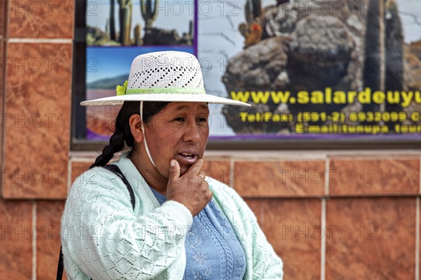 Woman in traditional dress wearing hat on the street in front of a tiled wall with advertising, people in an altiplano near Uyuni in Bolivia