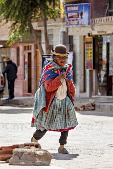Elderly woman in traditional clothes with melon and colorful scarf walks on cobblestone street, people in Altiplano near Uyuni in Bolivia