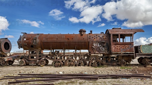 Rusted locomotive under a cloudy blue sky with graffiti along the body, old rusty locomotives at the train cemetery near Uyuni in the Altiplano in Bolivia