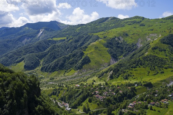 Lush green mountains with scattered village under blue sky and clouds, viewpoint near Gudauri, view of the mountainous landscape around Mleta, Mtskheta-Mtianeti region, Georgian Military Highway, High Caucasus, Georgia