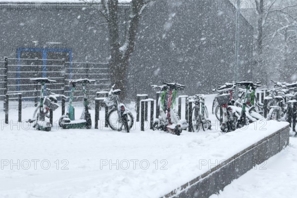 Tailored parked e-scooters and bicycles in winter during snowfall with snowflakes in the picture, Germany