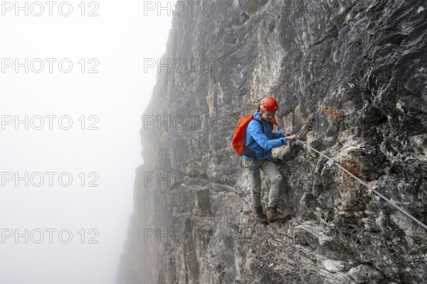 Mountaineers on a steep rock face on the Via Ferrata Oliva Detassis via ferrata in fog, steep mountains covered in clouds, Brenta Mountains, Trentino, Italy