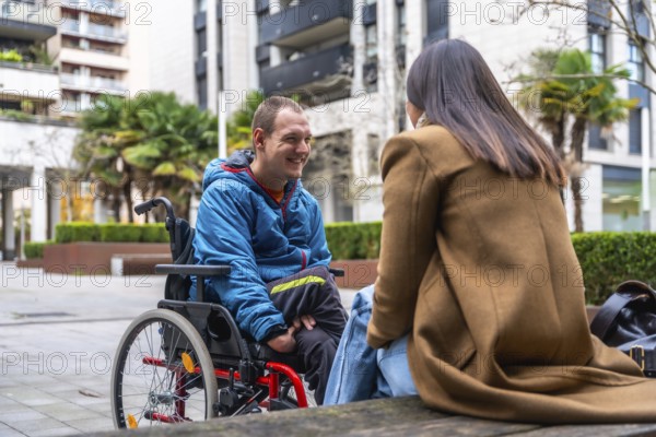 Young man in a wheelchair smiling and talking with a woman outdoors in an urban setting, representing concepts of diversity, inclusion, communication, and accessible lifestyle