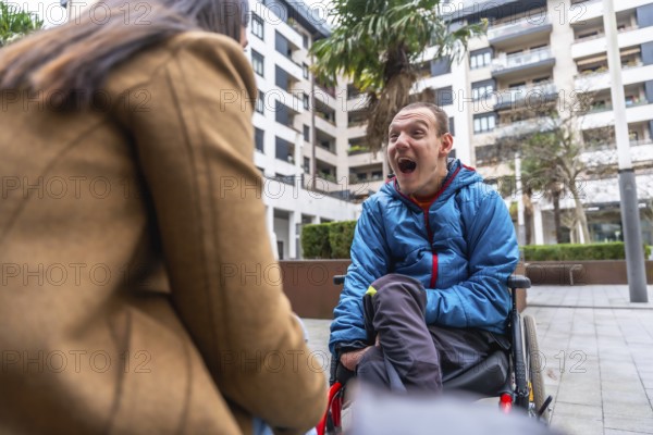 Disabled young man sitting in a wheelchair expressing joy and communicating with a caregiver or friend in an urban setting, representing inclusion and support