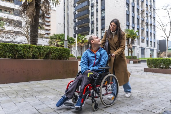 Woman caregiver is pushing a young man in a wheelchair on a paved outdoor area, both smiling and interacting, symbolizing support, companionship, and accessibility in daily life