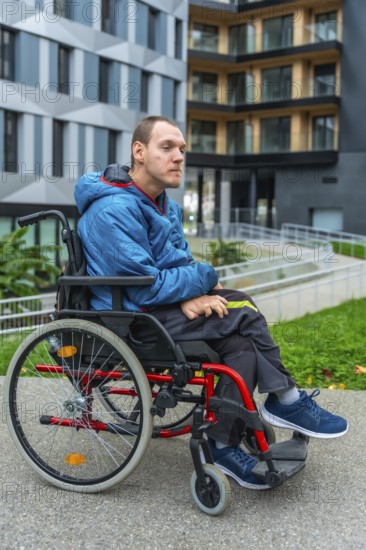 Young man living with a disability sitting in a manual wheelchair on a paved path outside modern urban buildings featuring an accessibility ramp, conveying themes of inclusion and independent living