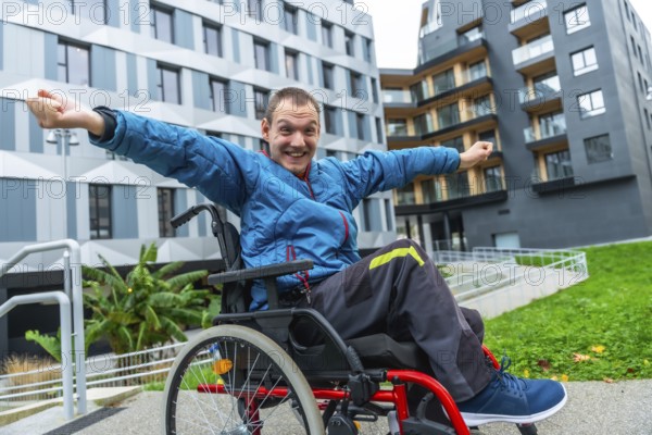 Smiling man in a wheelchair with arms outstretched expressing joy and freedom while navigating an accessible urban environment with modern buildings and green spaces