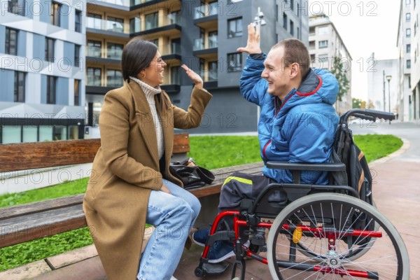Young man with a physical disability sitting in a wheelchair and celebrating with a high five, interacting with a supportive female friend or caregiver on a bench in an urban setting