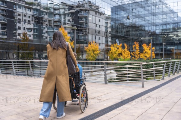 Woman in a brown coat pushing a young person in a wheelchair along a modern urban sidewalk, navigating the city street with glass buildings and autumn trees