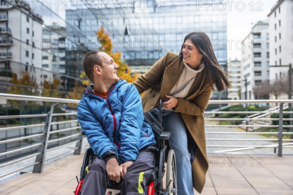 Young man in a wheelchair smiling and chatting with a supportive woman on a sunny urban street, enjoying inclusive companionship, mobility and joyful independence together