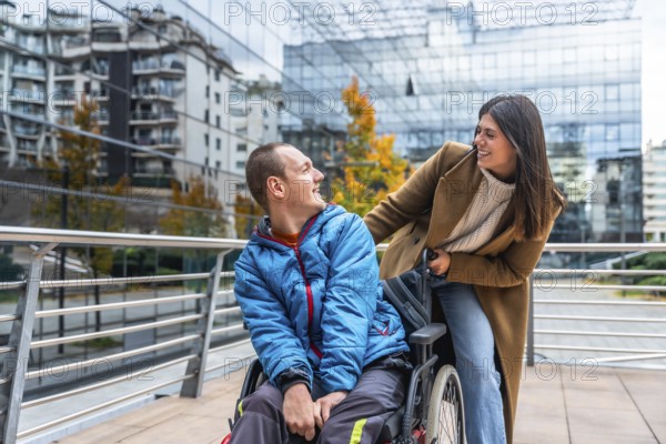Caregiver woman pushes smiling young man in wheelchair along a city sidewalk, sharing a warm moment of support, inclusion and friendship amid urban architecture and trees