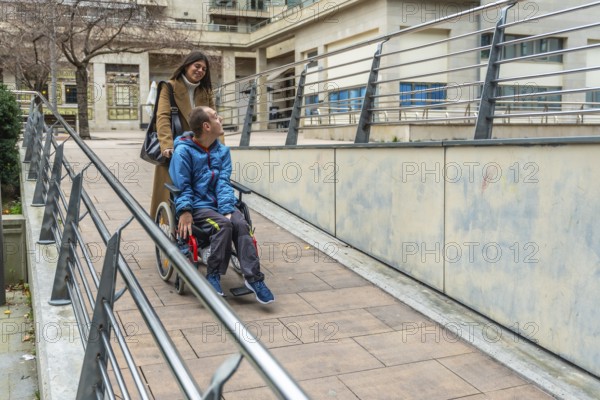 Woman pushing a young man with a disability in a wheelchair, navigating an accessible ramp with railings in an urban setting, receiving support and care