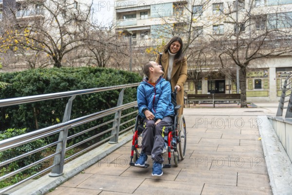 Caretaker pushing young man in wheelchair along accessible urban path, both smiling and sharing a warm moment of friendship, independence and inclusive outdoor mobility