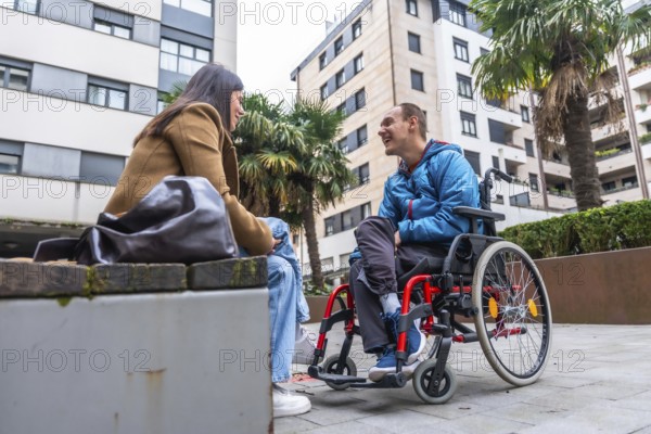 Young man with disability in a wheelchair discussing and smiling with a caring woman sitting on a bench in an urban setting. Representing concepts of inclusion. Friendship. And positive communication
