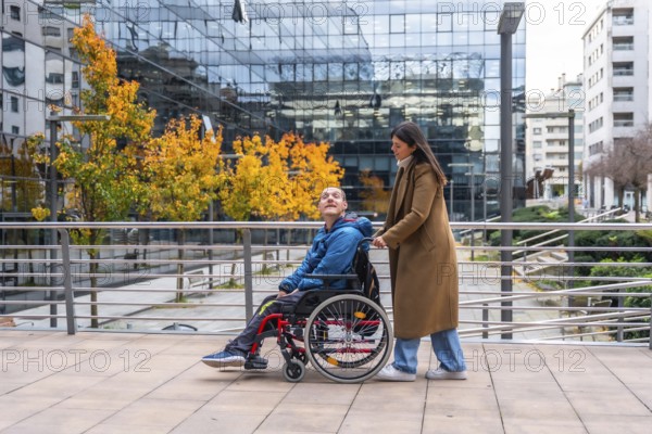 Woman pushing a smiling man in a wheelchair along an urban sidewalk during autumn, showcasing support, companionship, and accessibility in a modern city environment