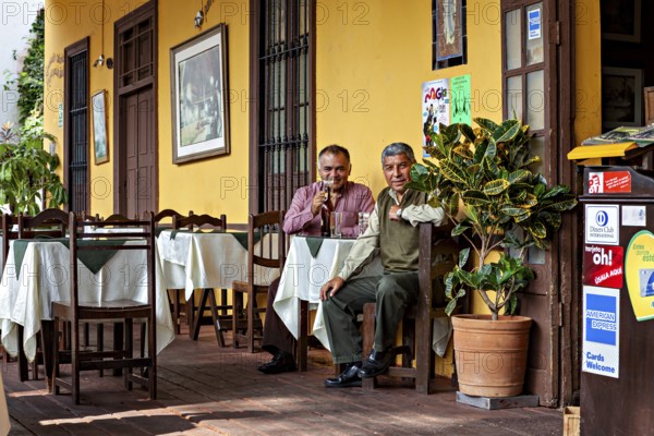 Two men sit comfortably outdoors at a table in front of a yellow building and enjoy the atmosphere, Two men in a restaurant in Lima in Peru