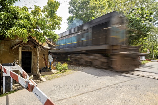 A fast-moving train passes a rural level crossing next to a small house, a level crossing with a barrier and a train in Sri Lanka