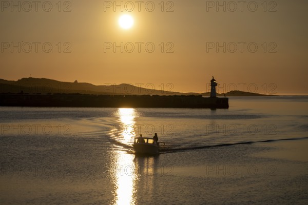 A small motorboat sails through the harbor entrance with lighthouse and midnight sun, Vardø, Finnmark, Norway