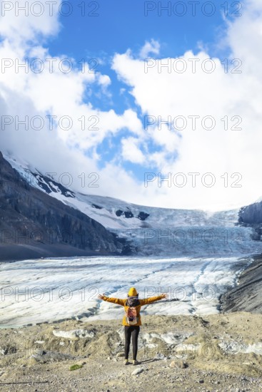 Female hiker with backpack and yellow jacket is enjoying the breathtaking view of the athabasca glacier in jasper national park, canadian rockies, alberta, under a beautiful cloudy sky