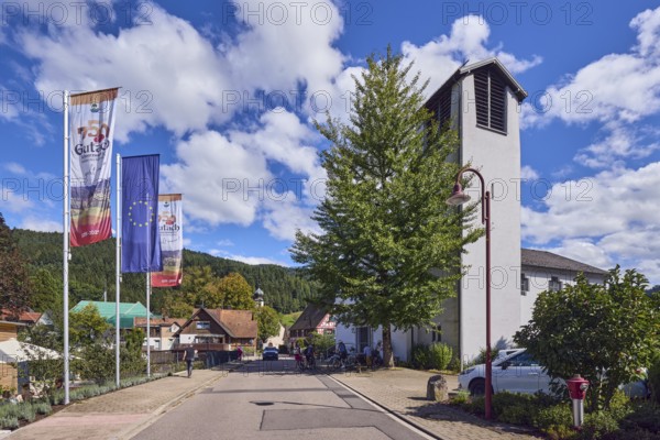 St. Peter and Paul parish church, church tower, flag on flagpole, European flag, 750 years Gutach Schwarzwaldbahn, the home of the Bollenhut, ... always goes, general architecture, Black Forest house, hill, hilly landscape with coniferous forest, road, pedestrians as accessories, blue sky, cumulus clouds, Kirchstraße, Gutach Schwarzwaldbahn, Black Forest, Ortenaukreis, Baden-Württemberg, Germany