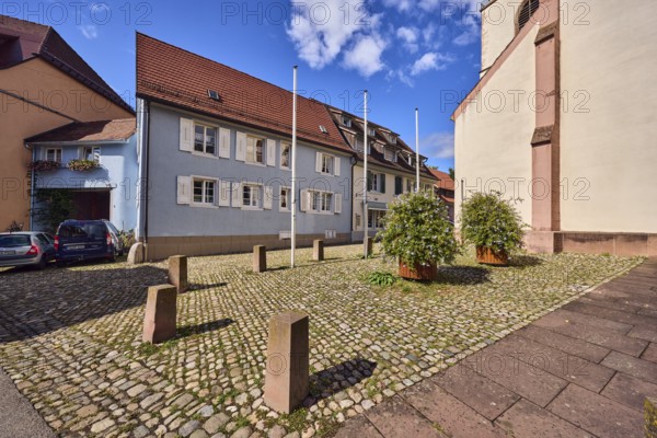 Houses, residential buildings, apartment blocks, church, facade with windows and shutters, dormers, flagpoles, planters with Cape Leadwort (Plumbago auriculata), bollards, cobblestone square, blue sky, cumulus clouds, Kirchstraße, Staufen im Breisgau, Black Forest, Breisgau-Hochschwarzwald district, Baden-Württemberg, Germany