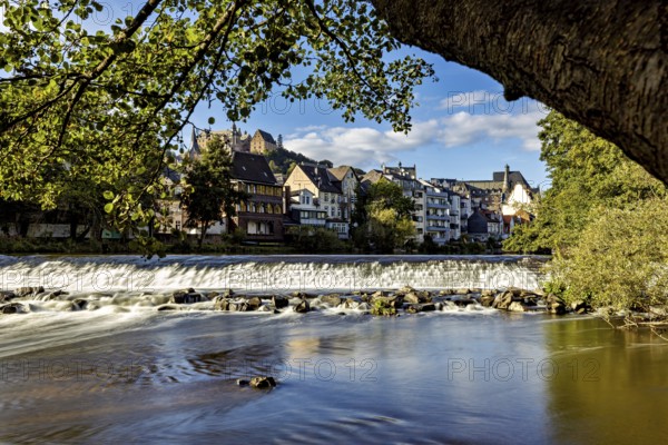 City scene with river and waterfall, surrounded by old buildings and trees under a blue sky, Das Wehr der Lahn in Marburg