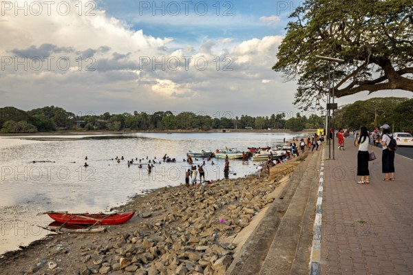 Boats on the riverbank with people on the promenade, rocky beach and cloudy sky in the background, people at a lake in Sri Lanka