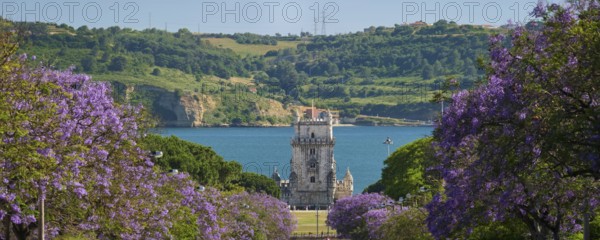 Scenic view of Belem Tower in Lisbon, Portugal, seen over a street with blooming purple jacaranda flower trees street with tourist sailboats on the Tagus River on sunset. Portugal