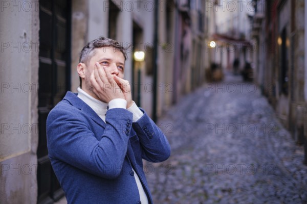 Man musician in a blue blazer and white sweater playing blues on a harmonica with eyes closed, standing outdoors in city street