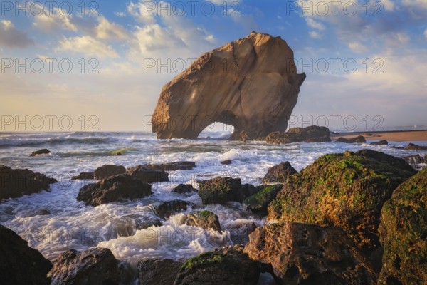 Penedo do Guincho, a large boulder rock arch at Praia da Santa Cruz, Portugal, with ocean waves and sandy beach on sunset