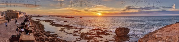 Panorama of a coastal promenade at sunset with rocks in the sea and a bright orange sky, Morocco, Essaouira