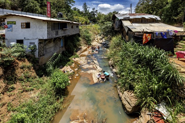 A river flows through a village, people wash clothes surrounded by nature and simple buildings, woman wash clothes in a river in the mountains near Kandy in Sri Lanka