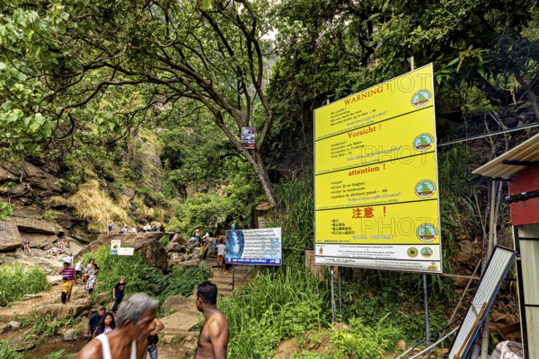 Trail in the jungle with warning signs and people surrounded by trees and rocks, tourists at the waterfalls in the mountains near Kandy in Sri Lanka